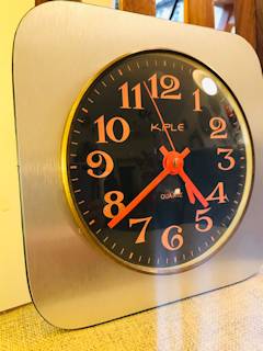 Horloge murale Kiple, support carré en bois teint en gris irisé, chiffres et aiguilles orange pop, mécanisme quartz.